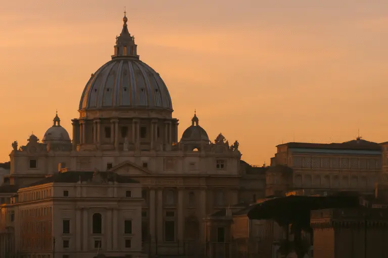 Basílica de São Pedro no Vaticano ao pôr do sol, com céu alaranjado ao fundo.